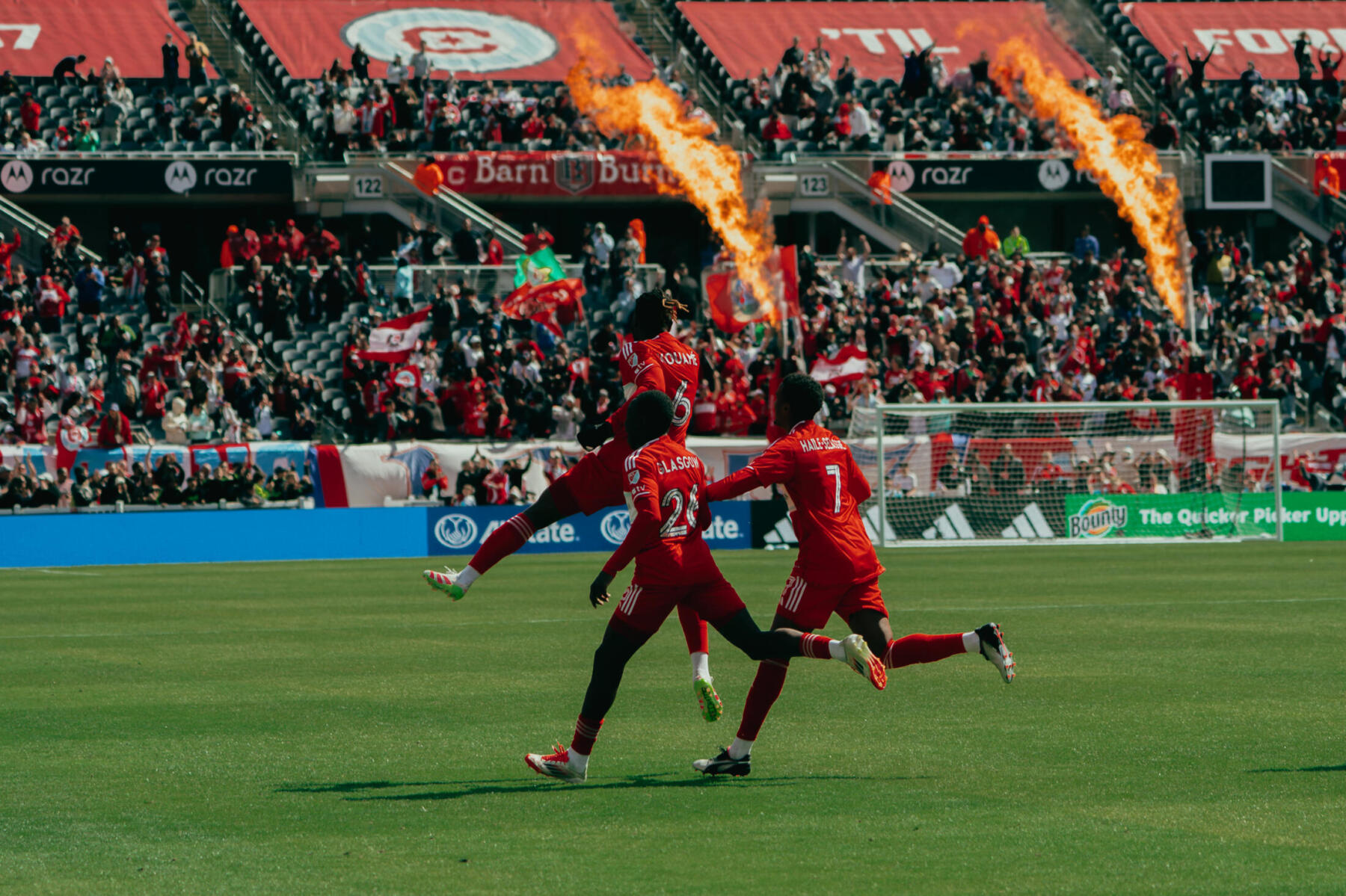 Chicago Fire players celebrate a goal with fire roaring in the background and fans in the crowd.