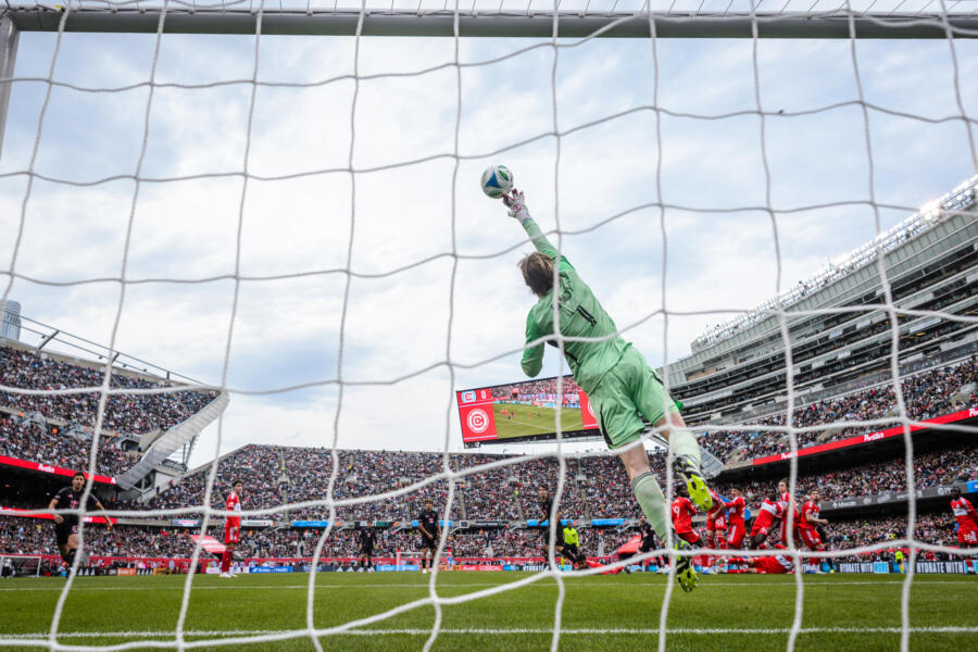 Chicago Fire goalkeeper jumps up and reaches to block a shot with his right hand.