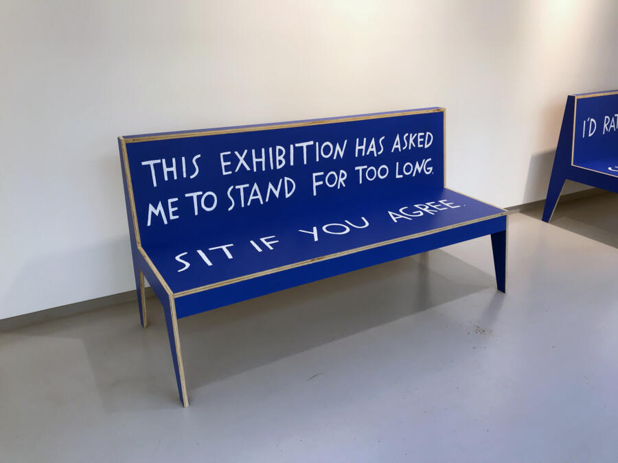 Blue wooden bench in a gallery with text painted on it.  The back of the bed bench, "This exhibit made me stand for too long." The seat reads, "Sit down if you agree."