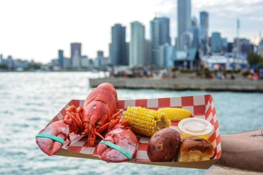 A lobster, potato, corn on the cob, and a roll held out in front of a lake.