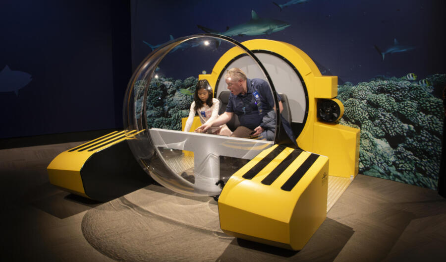 Visitors sit inside a model of a Triton Submersible, where they visualize an underwater expedition