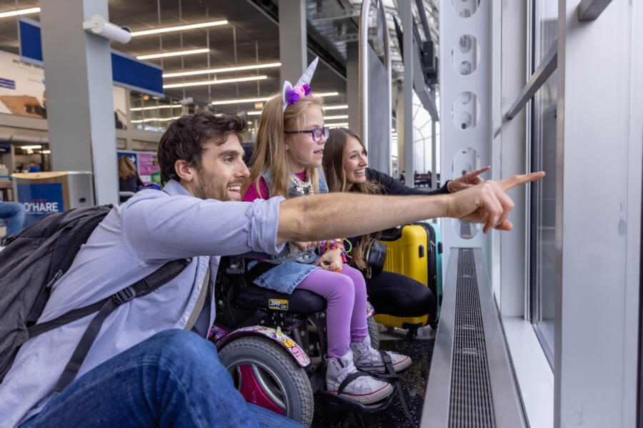 A child in a wheelchair looks out the airports window at planes with her parents by her side