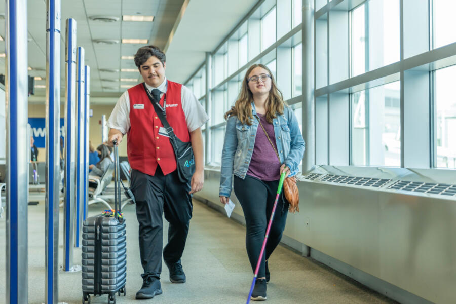 A woman walking with a cane is escorted by an airport staff member rolling her suitcase.