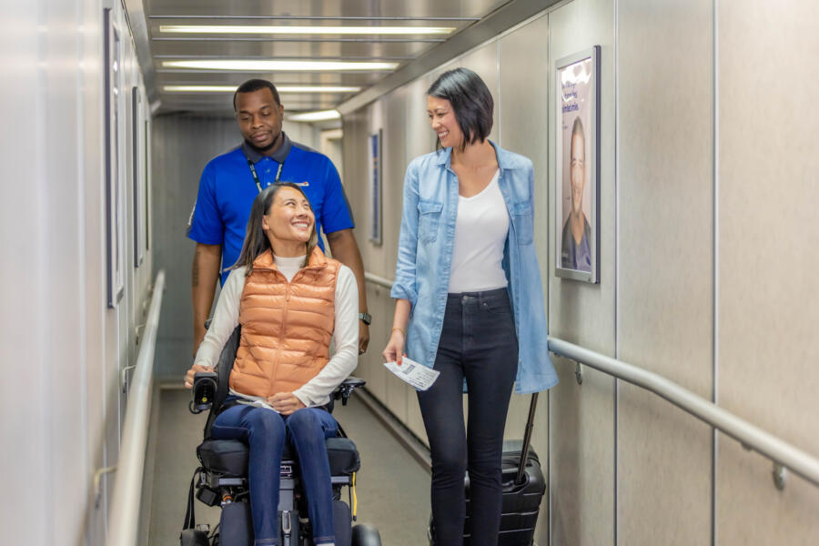 A woman in a wheelchair on the jet bridge entering an airplane with a friend and an airport staff member by her sides.