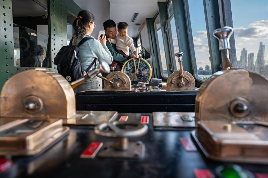 A father holds a toddler at the helm of a historic ship's wheelhouse, with brass controls in the foreground and a woman taking a photo.