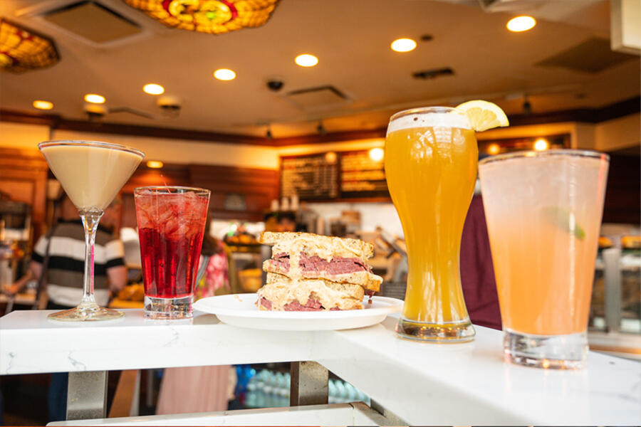 A marble counter displays a variety of drinks including a martini, a red iced beverage, a tall glass of beer with a lemon slice, and a pink cocktail, alongside a Reuben sandwich, set in a warm, vintage-style restaurant interior.