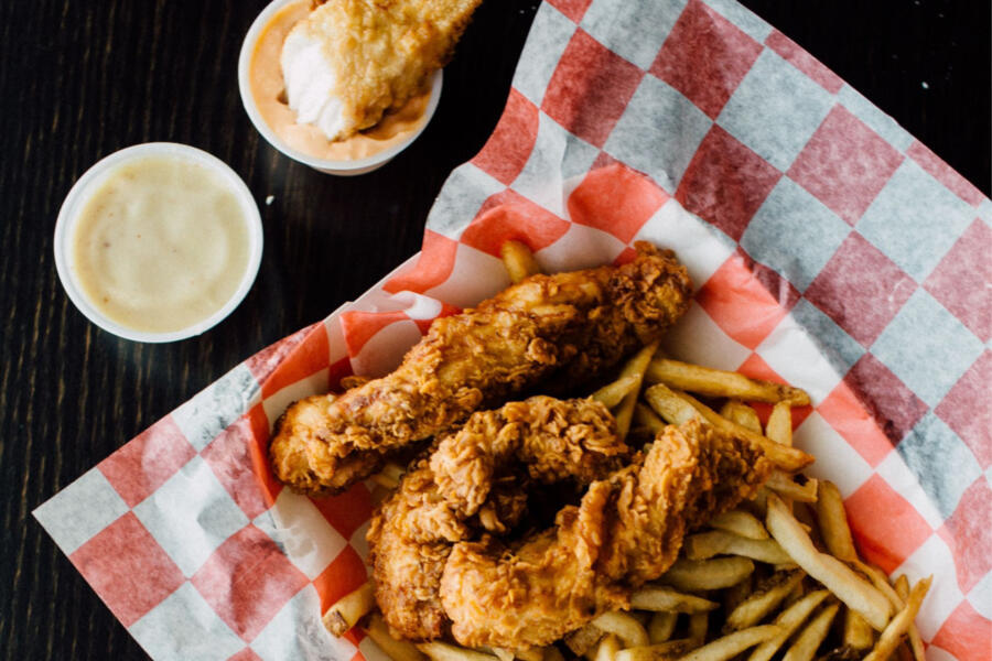 A serving of crispy fried chicken tenders and golden French fries on red and white checkered paper, accompanied by two dipping sauces, one with an extra chicken tender partially dipped.