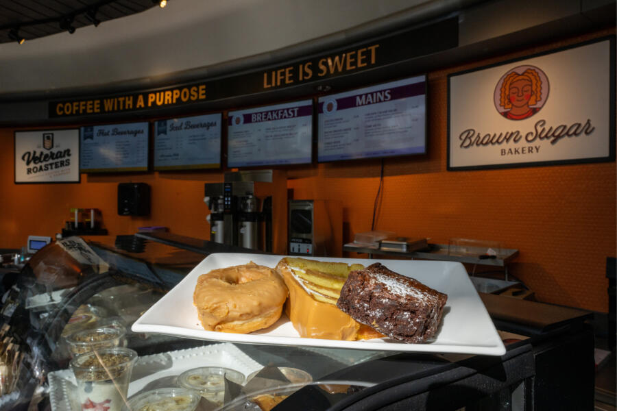 A white plate holds a maple-glazed donut, a slice of caramel cake, and a chocolate brownie, displayed on a counter in front of Brown Sugar Bakery with digital menu boards and coffee machines in the background.