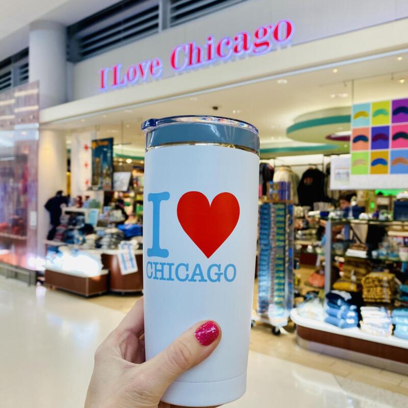 A travel mug with "I Heart Chicago" written on it being held up in front of the I love Chicago store at O'Hare Airport