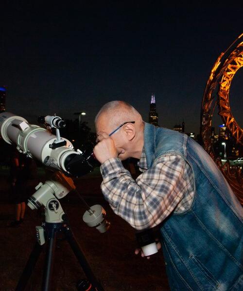 Man looking through a telescope at night with the Chicago skyline and a lit spiral sculpture behind him.