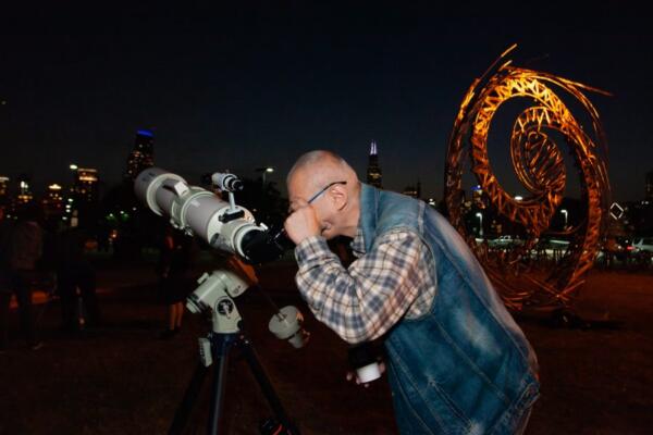 Man looking through a telescope at night with the Chicago skyline and a lit spiral sculpture behind him.