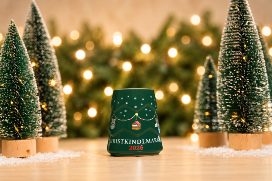 The green Christkindlmarket Non-alcoholic Tree Mug 2026 placed next to small Christmas trees.